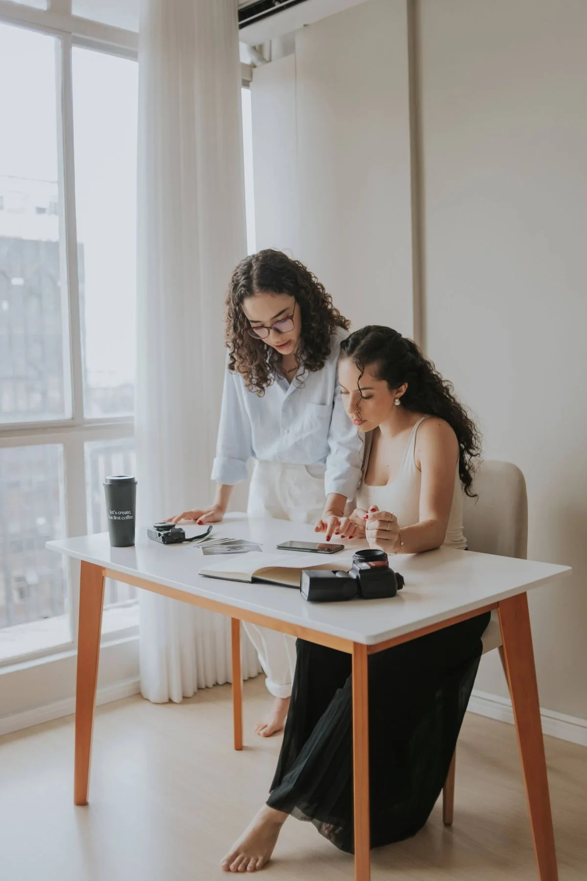 Two women reviewing creative work together beside a large window.