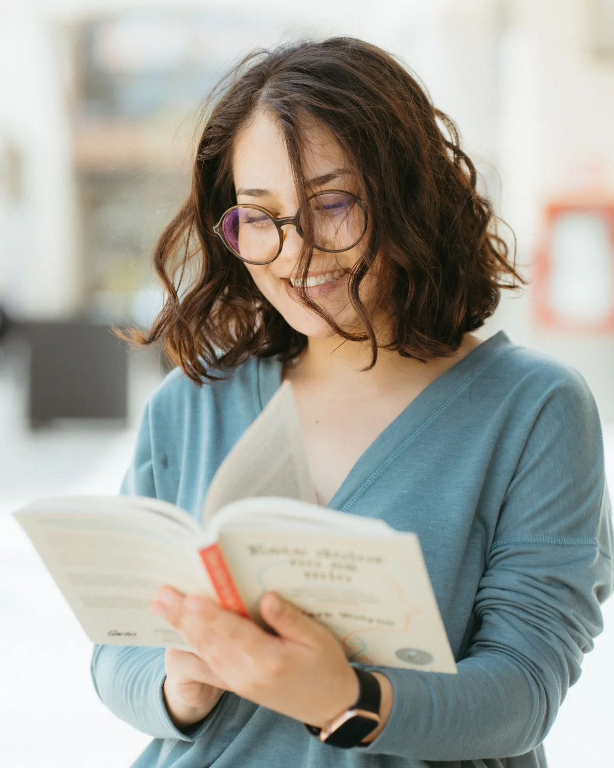 Woman in glasses reading a book in a bright room.