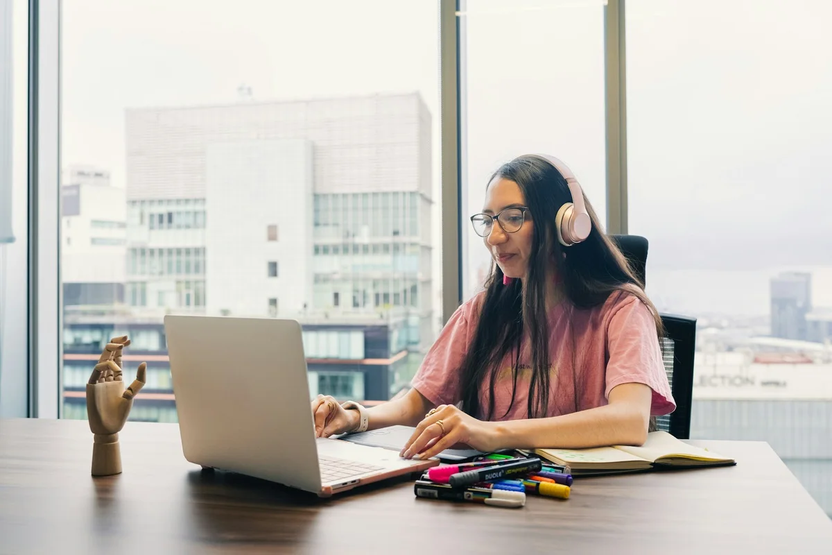 Woman wearing headphones working on a laptop beside a bright studio window.
