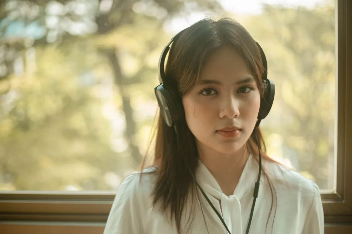 Young woman wearing headphones beside a daylight-filled window.