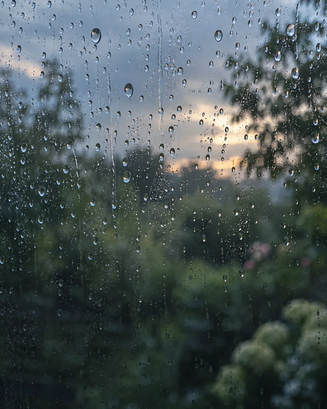 Raindrops run down a window with a garden softly out of focus beyond it.