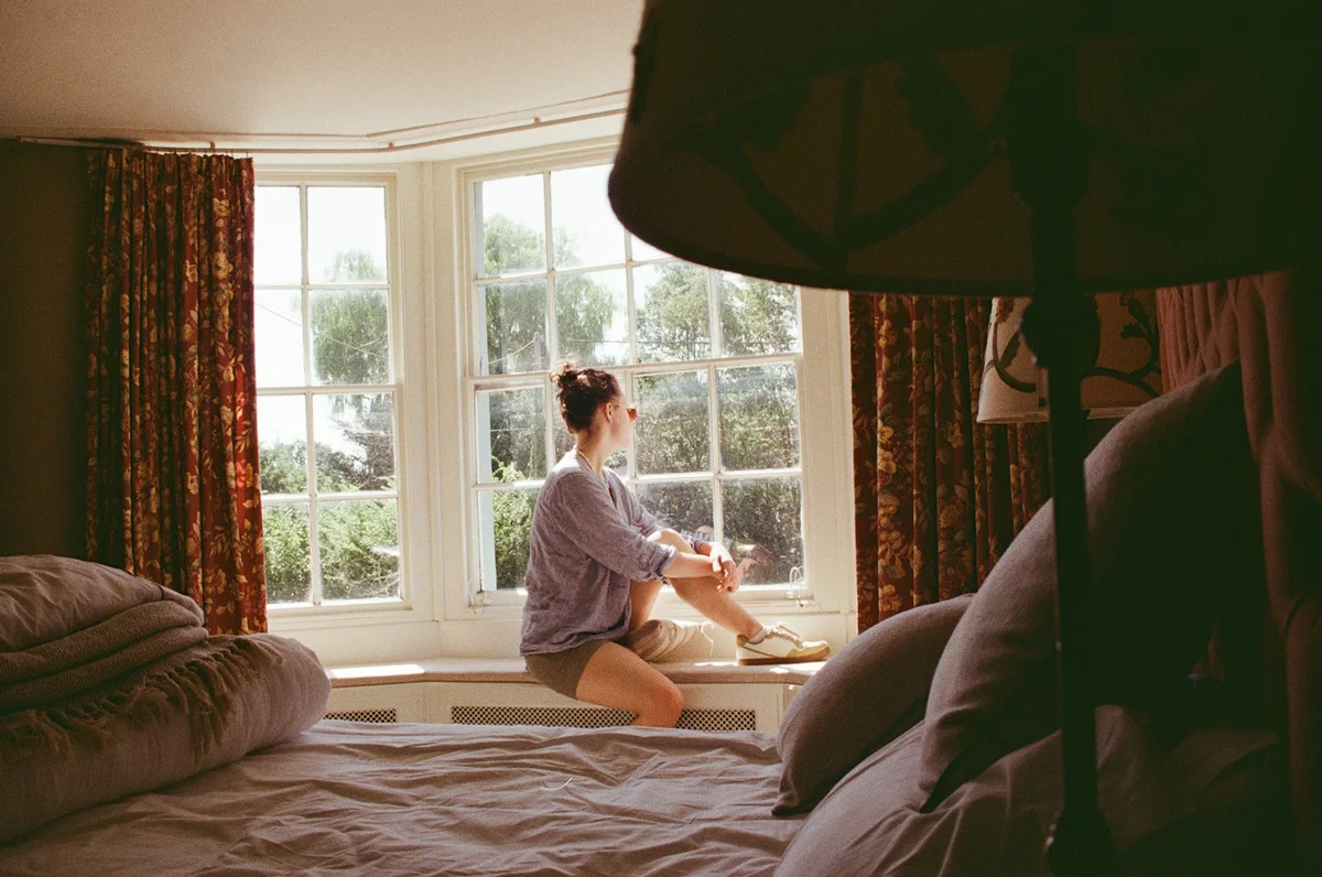 A woman sits at a bay window, looking outside.