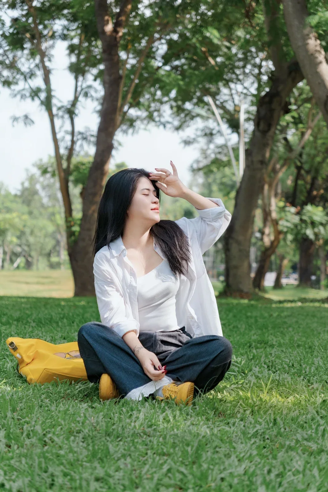 Woman sitting cross-legged on grass beneath trees, looking into the daylight.