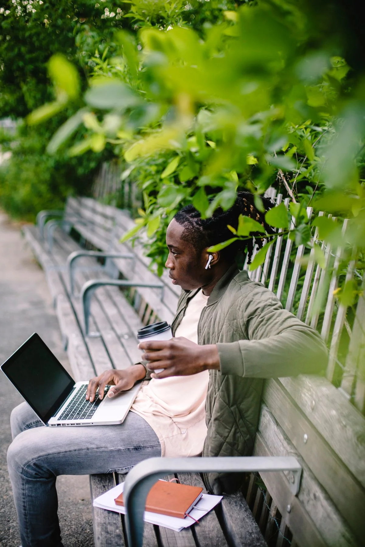Man pausing with a coffee on a park bench, with a laptop and notebooks beside him.