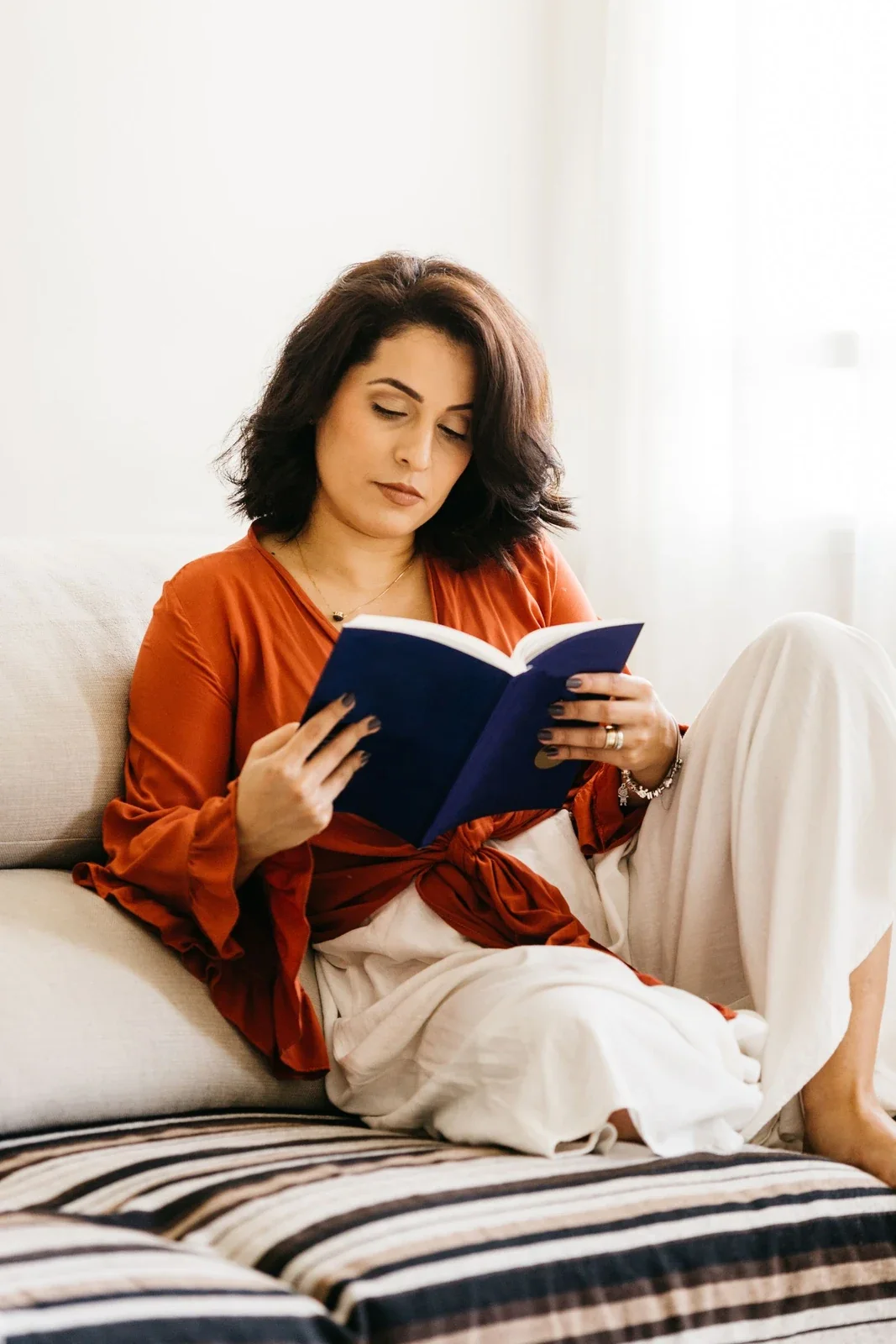 Woman reading a blue book on a pale sofa in a bright room.